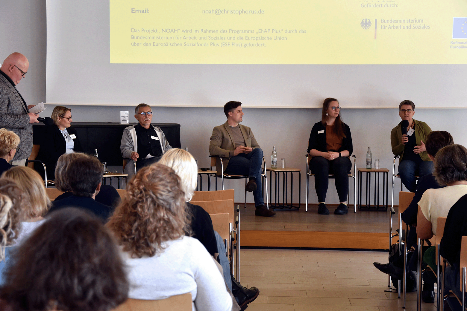 Auf einer Podiumsdiskussion sieht man fünf Personen auf einer niedrigen Bühnen sitzend im Gespräch mit dem Publikum vertieft. Im Hintergrund ist eine große Präsentation auf dem Beamer eingeblendet.