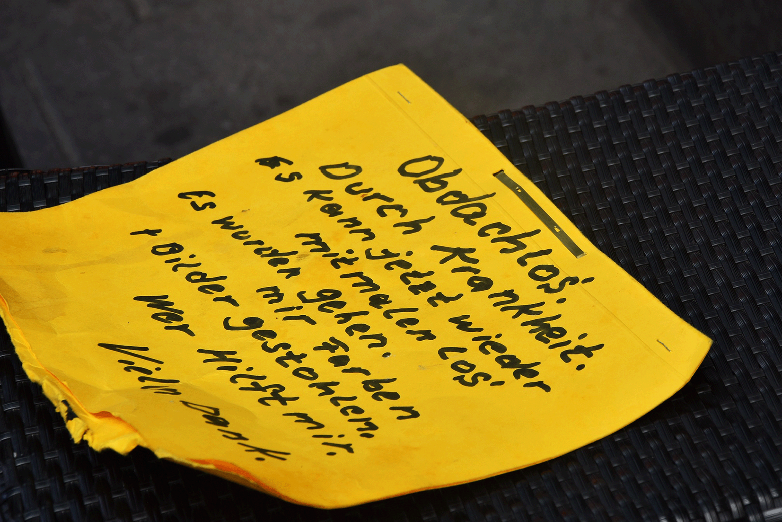 Auf einem gelben Notizzettel wurde mit schwarzem Filzstift notiert: "Obdachlos durch Krankheit. Es kann jetzt wieder mit malen losgehe. Es wurden mir Farben + Bilder gestohlen. Wer hilft mir? Vielen Dank."